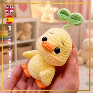 Puede incluir: Un patito amarillo hecho a mano con ganchillo con pico y patas naranjas, y ojos negros. El patito tiene un brote verde en la cabeza. El texto "Pattern in English & Español" es visible en el lado izquierdo de la imagen.