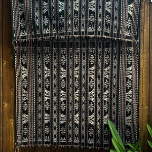 May include: A patterned textile in black and white, featuring vertical stripes and decorative elements. The fabric has a fringed edge and is displayed against a wooden backdrop. The design incorporates brown and white accents, creating a striking contrast.