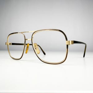 May include: Gold-rimmed eyeglasses with a double bridge and black temples. The lenses are clear, and the frames have a rectangular shape. The glasses have a vintage aesthetic.