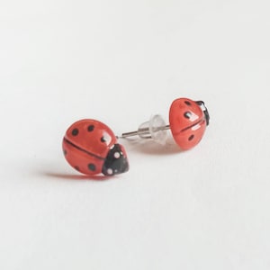 Op de afbeelding: Een paar oorbel studs in de vorm van een lieveheersbeestje. De oorbel studs zijn rood met zwarte stippen en hebben zilveren pinnen.