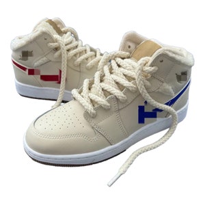 May include: Cream-colored high-top sneakers with a white sole and a blue and red logo. The laces are white and the shoes have a fuzzy lining.