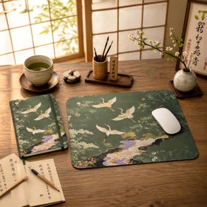 May include: A rectangular desk mat and a matching notebook, both featuring a dark green background with white crane illustrations, gold accents, and purple floral patterns. A white computer mouse rests on the mat, alongside a ceramic tea cup, a bamboo pen holder with brushes, and an open book with black text.