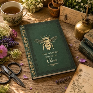 May include: A dark green hardcover journal with a gold foil bee illustration on the front cover. The text reads THE GARDEN NOTES OF Clara. The spine features a gold leaf pattern. The book is surrounded by a teacup, scissors, and botanical cards.