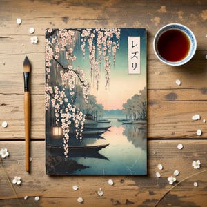 Op de afbeelding: Een notitieboek met een Japanse landschapsillustratie met een rivier, boten en kersenbloesems. De kaft heeft Japanse tekst. Een penseel en een kopje thee liggen naast het notitieboek op een houten oppervlak.