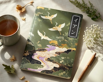 May include: A green journal with a crane design and Japanese text. The cover features three cranes in flight over a landscape with gold and purple accents. A cup of tea, a pen, and a small notebook are also visible.