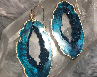 May include: Teal blue agate earrings with gold-toned metal settings. The earrings are shaped like a geode with a clear center.