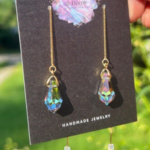 May include: A pair of gold chain earrings with iridescent teardrop crystals hanging from them. The earrings are displayed on a black card with the text "KG Design & Decor by KARIC GLOVER" and "HANDMADE JEWELRY".