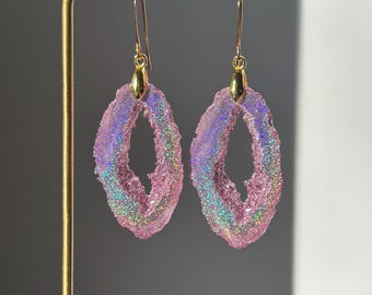 May include: A pair of drop earrings featuring oval-shaped, translucent pink resin pendants with a textured, crystalline surface that displays a holographic, iridescent shimmer. The pendants are suspended from gold-toned metal hooks.