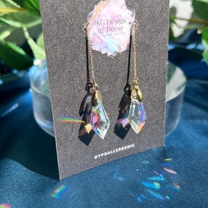 May include: A pair of gold-tone chain earrings featuring iridescent, diamond-shaped crystal pendants. The earrings are displayed on a dark gray card labeled KG Design & Decor and Hypoallergenic. The crystals reflect light, creating small rainbow prisms.