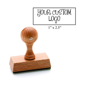 May include: Wooden self-inking stamp with a 1 inch by 2.5 inch rectangular imprint area. The stamp has a wooden handle and a rectangular base. The text "YOUR CUSTOM LOGO" is printed on the stamp.