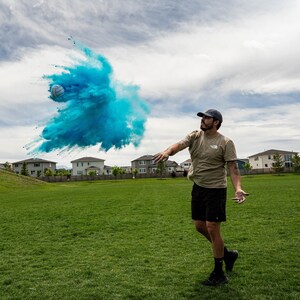 以下が含まれることがあります： 人が白いボールを空中に投げ、青い粉の爆発を起こしています。その人は茶色のTシャツ、黒いショートパンツ、黒い野球帽をかぶっています。