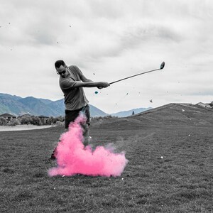 May include: A person wearing black and white clothing swings a golf club, producing a pink cloud of smoke behind them.