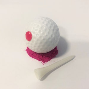 May include: A white golf ball with a red dot on it, sitting on a bed of pink sand. A white plastic golf tee is lying on the white surface.
