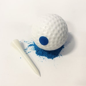 May include: A white golf ball with a blue dot on it, sitting on a pile of blue powder. A white golf tee is lying next to the ball.