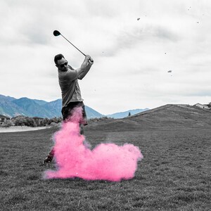May include: A golfer swings a club, creating a pink cloud of smoke behind him. The golfer is wearing a grey shirt and black shorts. The background is a golf course with green grass and a mountain range in the distance.