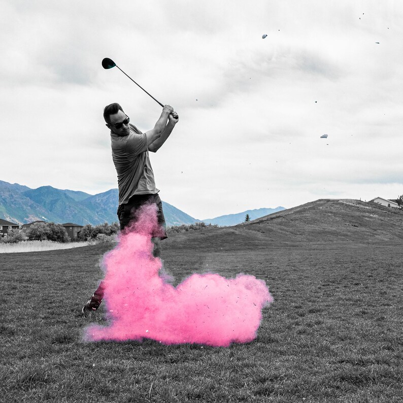 May include: A golfer swings a club, creating a pink cloud of smoke behind him. The golfer is wearing a grey shirt and black shorts. The background is a golf course with green grass and a mountain range in the distance.