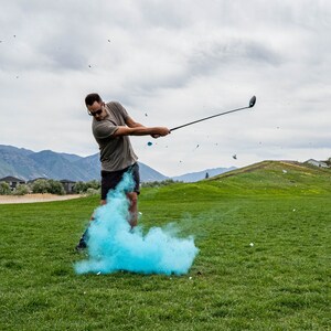 May include: A person swings a golf club, creating a blue cloud of smoke. The person is wearing a grey shirt and black shorts. The background is a green grassy field with a mountain range in the distance.