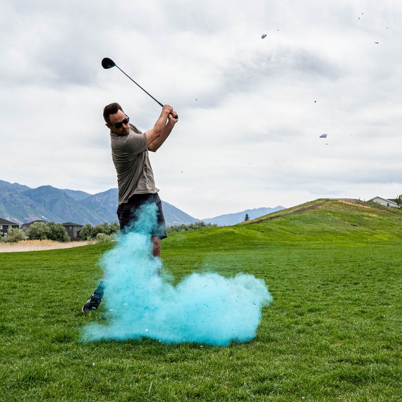 May include: A person swings a golf club, creating a blue cloud of powder behind them on a green golf course.