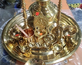 May include: A set of brass puja items, including a large pot, two oil lamps, a small bowl, and several smaller bowls, all arranged on a large brass tray. The items are all gold in color and have intricate designs.