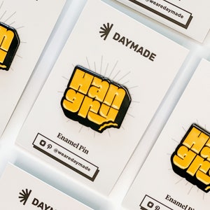 May include: A yellow and black enamel pin with the text "Hangry" in a bold, blocky font. The pin is on a white card with the text "DAYMADE" and "Enamel Pin" in black text.