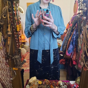 Könnte beinhalten: Eine teal-farbene Bluse mit vertikalen Streifen und Bindedetail, kombiniert mit einem schwarzen Rock. Die Bluse hat lange Ärmel und eine Knopfleiste. Der Spiegel spiegelt verschiedenen Schmuck und Accessoires wider, was eine Vintage-Ästhetik erzeugt.