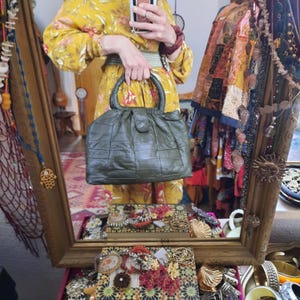 Könnte beinhalten: Olivgrüne, gesteppte Handtasche mit rundem oberem Griff und Klappenverschluss. Die Tasche hat ein strukturiertes Design mit einem Gittermuster. Die Tasche wird vor einem Spiegel gehalten, der ein gelbes Blumenkleid reflektiert.