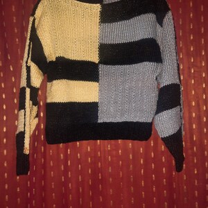 Pu&ograve; includere: Un maglione a blocchi di colore con scollo a barchetta. Il maglione &egrave; diviso in quattro sezioni: beige, nero, grigio e nero. Le maniche sono lunghe e presentano un motivo a scacchi nero e beige su una manica e strisce nere e grigie sull'altra.