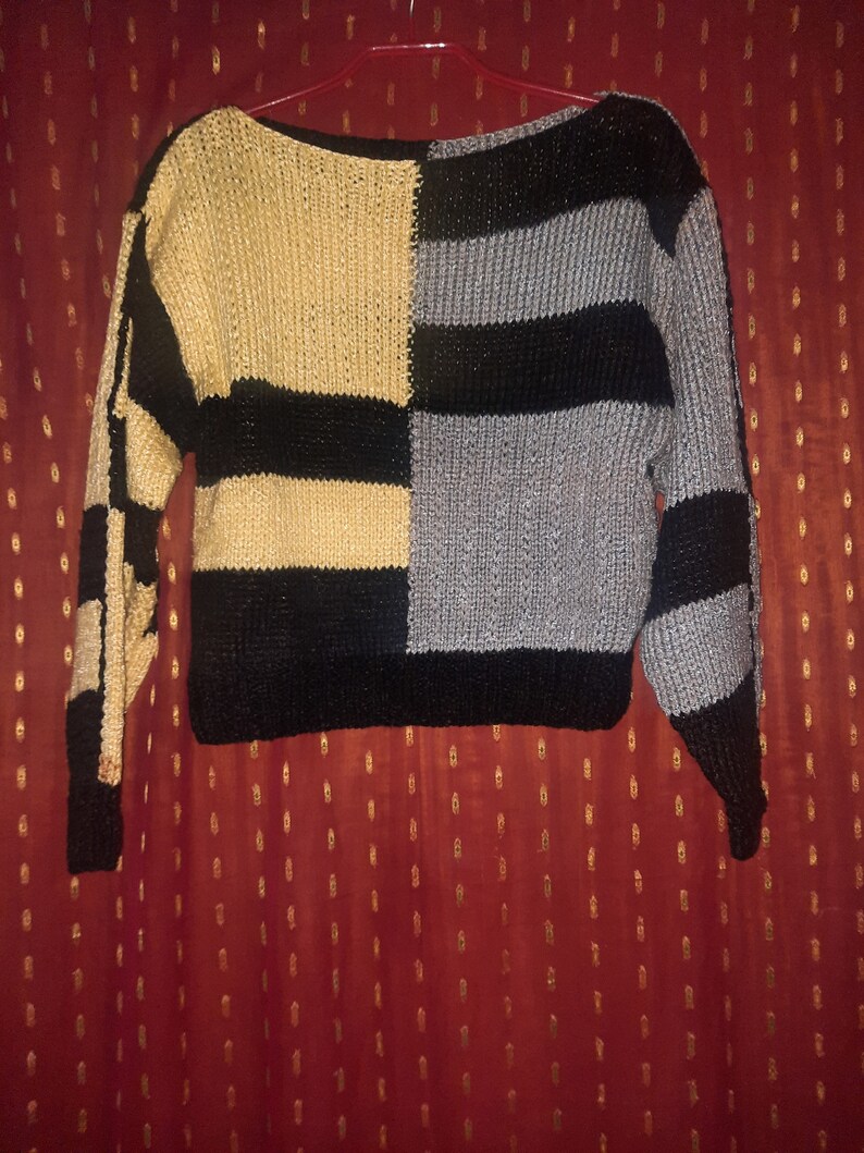 Pu&ograve; includere: Un maglione a blocchi di colore con scollo a barchetta. Il maglione &egrave; diviso in quattro sezioni: beige, nero, grigio e nero. Le maniche sono lunghe e presentano un motivo a scacchi nero e beige su una manica e strisce nere e grigie sull'altra.