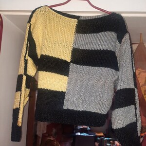 Pu&ograve; includere: Un maglione a blocchi di colore con un design unico. Il maglione presenta una combinazione di pannelli oro, neri e grigi. Le maniche lunghe e il corpo sono divisi in sezioni geometriche. Il maglione &egrave; appeso a una gruccia rossa.