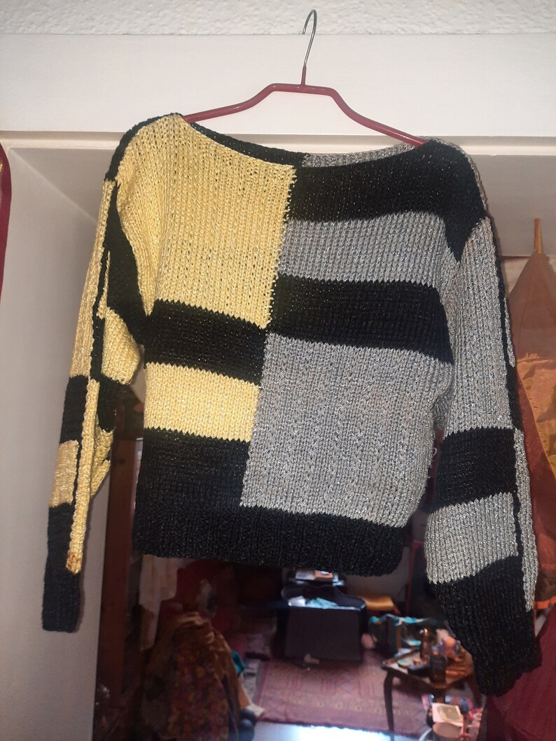Pu&ograve; includere: Un maglione a blocchi di colore con un design unico. Il maglione presenta una combinazione di pannelli oro, neri e grigi. Le maniche lunghe e il corpo sono divisi in sezioni geometriche. Il maglione &egrave; appeso a una gruccia rossa.