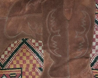 May include: Brown suede cowboy boots with a pointed toe and a low heel. The boots have decorative stitching and are set against a patterned background.