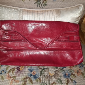 Könnte beinhalten: Eine rote Leder-Clutch mit Klappenverschluss und einem dekorativen Riemen.