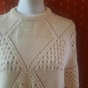 Könnte beinhalten: Ein cremefarbener Häkelpullover mit Rautenmuster. Der Pullover hat einen runden Ausschnitt und kurze Ärmel.