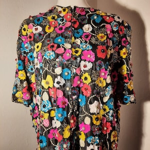 Pode incluir: Casaco preto de manga curta adornado com bordados florais coloridos. As flores são em tons de rosa, amarelo, azul e branco, com detalhes de contas. O casaco é exibido em um manequim, mostrando o design.