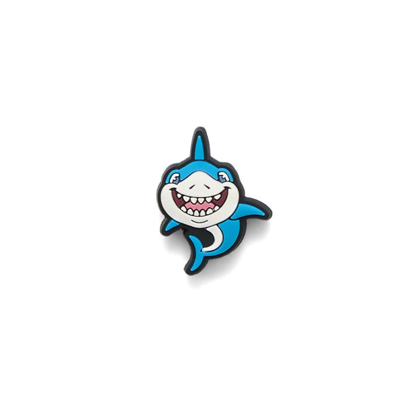 May include: A cartoon-style shark charm in shades of blue, white, and black. The shark has a wide, toothy grin and is smiling. The charm is likely made of a rubbery material and is designed to be a decorative accessory.