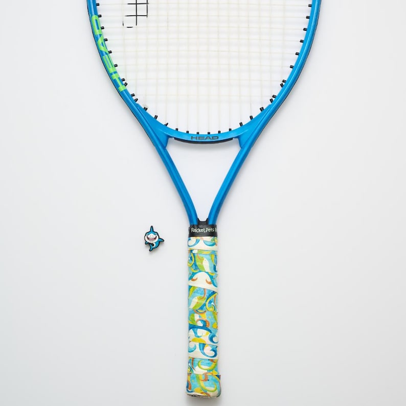 May include: A blue tennis racket with a white string net. The handle is wrapped in a colorful patterned grip. A small shark-shaped charm is placed near the handle. The word "HEAD" is visible on the racket.