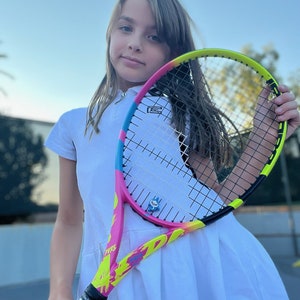 May include: A colorful tennis racket with a pink, yellow, and blue design. The racket has the text "PURE" and "Racket Pets" on it. The grip is decorated with a blue and white pattern. The racket is held by a person wearing a white dress.