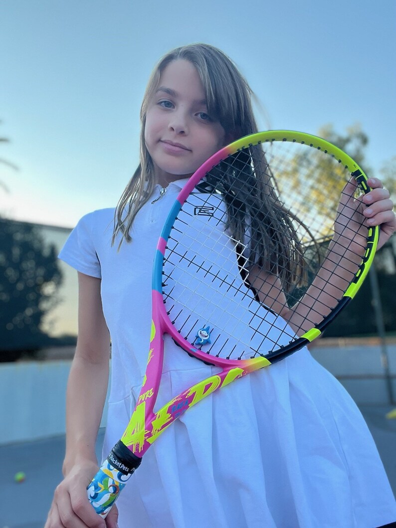 May include: A colorful tennis racket with a pink, yellow, and blue design. The racket has the text "PURE" and "Racket Pets" on it. The grip is decorated with a blue and white pattern. The racket is held by a person wearing a white dress.