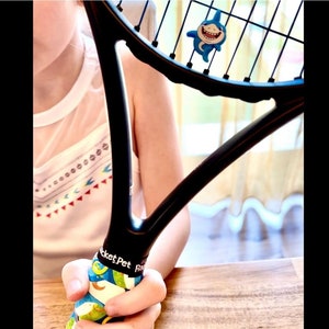 May include: Black tennis racket with a blue and white shark-shaped charm attached to the strings. The handle is wrapped in a colorful grip with the text "Rocket Pet". The racket is held by a person wearing a white shirt.