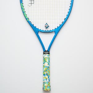 May include: A blue tennis racket with a shark logo and a patterned grip. The racket has a blue frame with green accents and the word "HEAD" printed on the head. The grip is wrapped in a colorful design with the words "RacketPets".