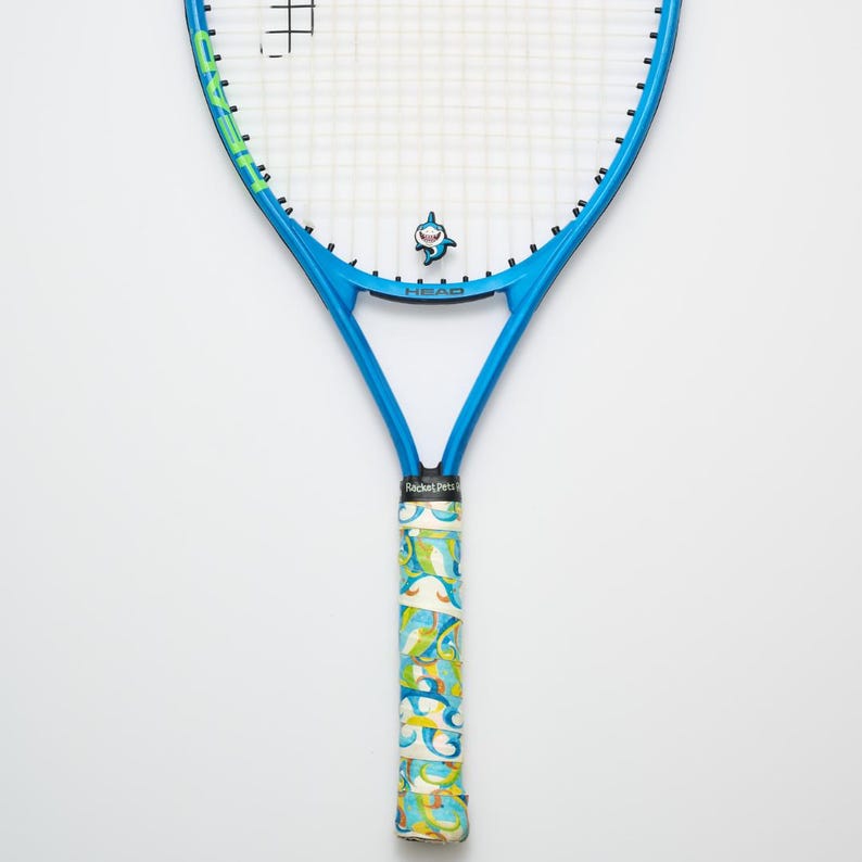 May include: A blue tennis racket with a shark logo and a patterned grip. The racket has a blue frame with green accents and the word "HEAD" printed on the head. The grip is wrapped in a colorful design with the words "RacketPets".