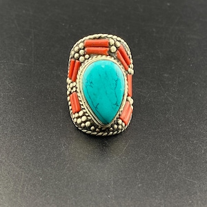 Puede incluir: Un anillo plateado con una gran piedra turquesa en forma de lágrima en el centro. El anillo está adornado con pequeños trozos de coral rojo y un borde de cuentas. El anillo está sobre un fondo oscuro.