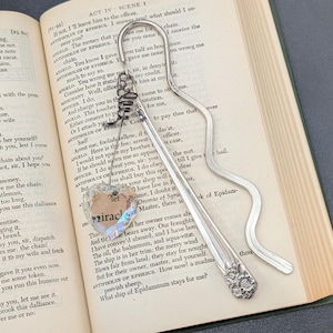 May include: A silver-toned metal bookmark with a wavy design and a floral pattern at the base. A wire-wrapped clear heart-shaped crystal charm hangs from the top, featuring the text "let miracles" printed on its surface, resting on an open book.
