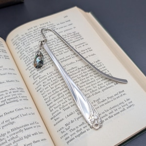 May include: A silver-toned metal bookmark featuring a curved handle with an embossed floral design at the base. A small, faceted, dark gray teardrop-shaped crystal charm hangs from a silver jump ring attached to the top of the bookmark.