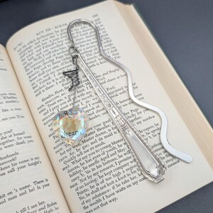 May include: A silver-toned metal bookmark featuring a wavy handle and a decorative flat end. A small silver charm of a bird is attached to the top, along with a faceted, iridescent heart-shaped crystal pendant that reflects light in multiple colors.