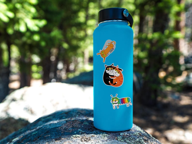 Op de afbeelding: Een turquoise waterfles met een zwarte dop en drie kleurrijke stickers. De stickers tonen cartoonkatten en een rups met een regenboogstreepontwerp. De fles staat op een rots.