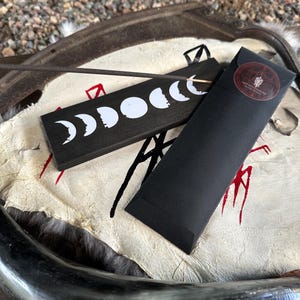 May include: A black incense holder with a white moon phase design and a black incense stick. A black paper sleeve with a red sticker is next to the holder. The items are on a white and tan surface.