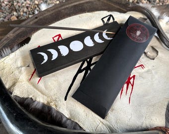 Puede incluir: Un porta incienso negro con un diseño de fases lunares blancas y un palito de incienso negro. Una funda de papel negro con una pegatina roja está al lado del soporte. Los artículos están sobre una superficie blanca y beige.