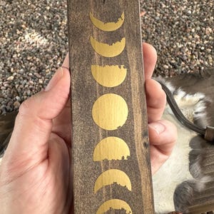 May include: A rectangular wooden plaque with a dark brown finish, featuring a series of gold-colored moon phase designs. The phases range from crescent to full moon. The plaque is held in a hand.