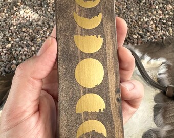Puede incluir: Placa rectangular de madera con acabado marrón oscuro, con diseños de fases lunares doradas. Las fases van desde la media luna hasta la luna llena. La placa se sostiene en una mano.
